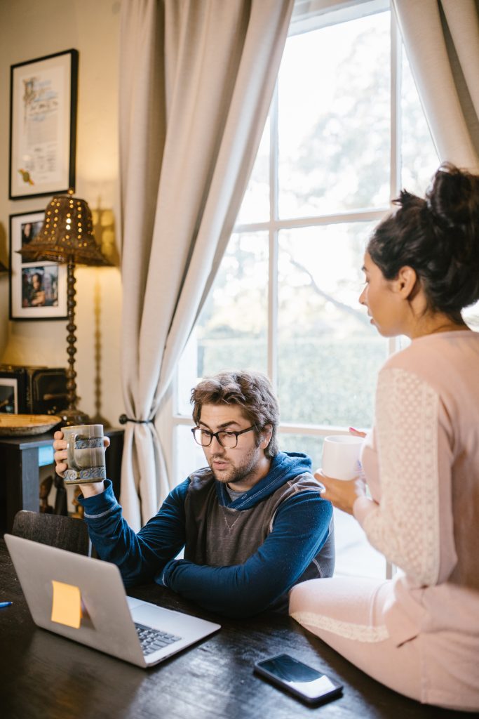man and woman holding mugs sitting in front of a laptop.